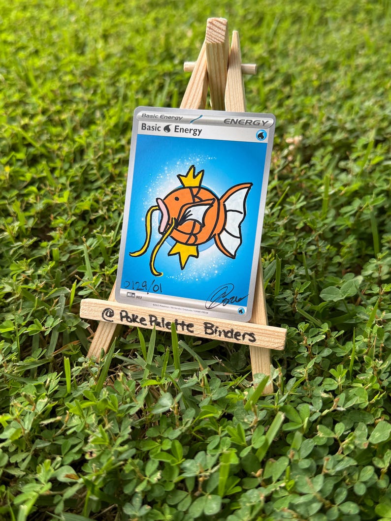 May include: A rectangular trading card featuring a blue background and a central orange fish illustration with yellow fins and whiskers. The text reads Basic Energy, Basic Water Energy, 2129/01, and a signature. The card rests on a small wooden easel.