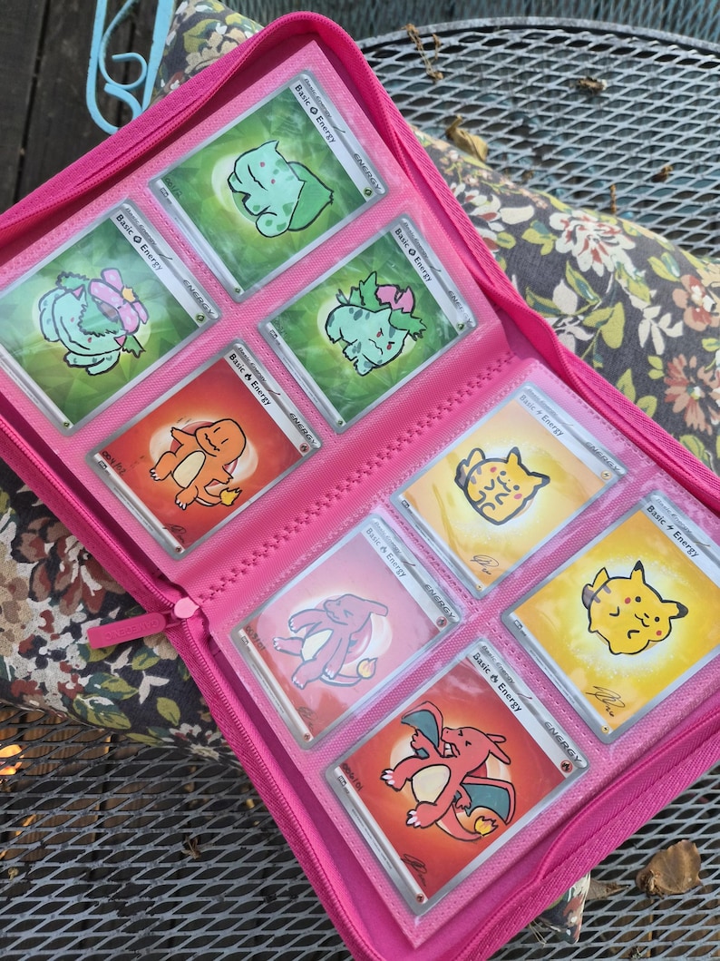 May include: A bright pink zippered trading card binder containing eight individual trading cards. The cards feature various illustrated characters including Bulbasaur, Charmander, Charizard, and Pikachu, each labelled as Basic Energy with a unique signature.