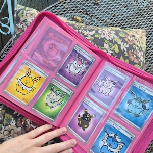 May include: A bright pink zipped binder containing multiple trading cards in clear plastic sleeves. The cards feature colourful illustrations of various creatures, including yellow, green, purple, and blue characters, each labelled as Basic Energy.
