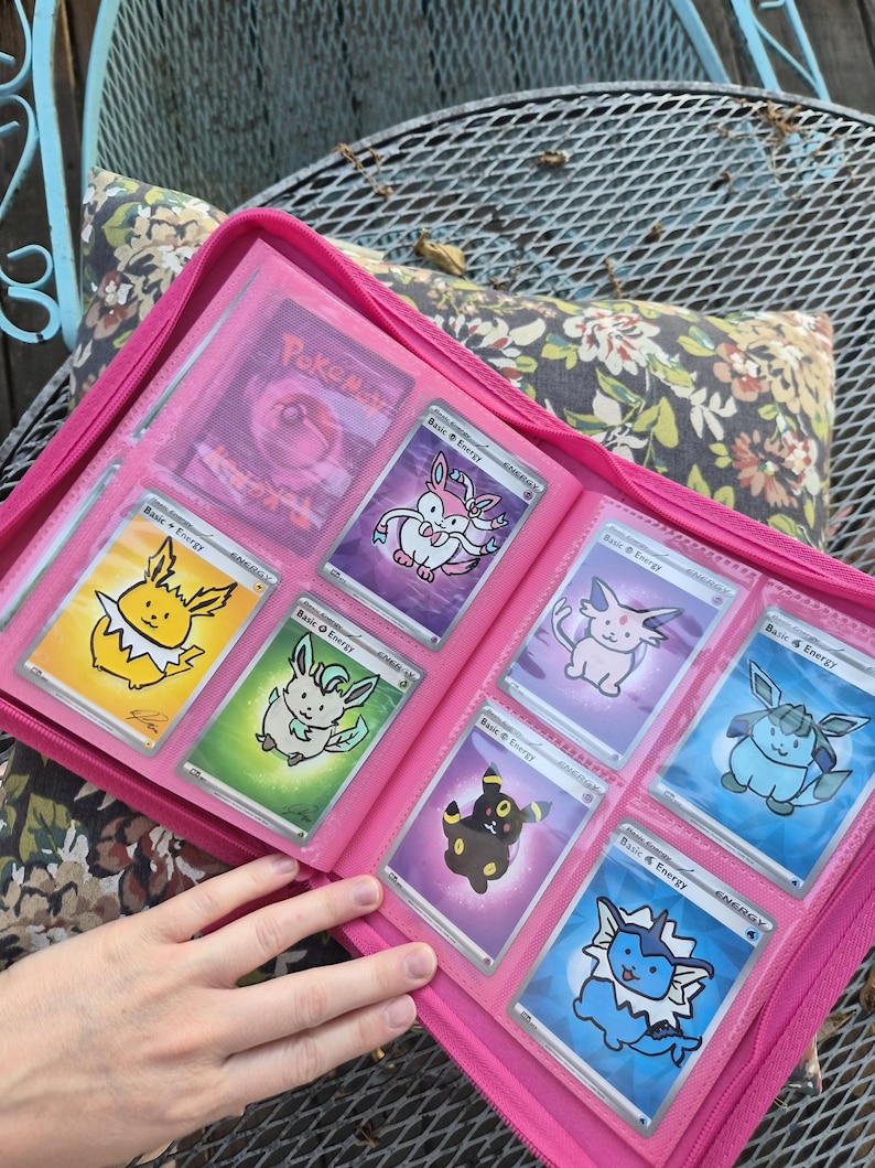 May include: A bright pink zipped binder containing multiple trading cards in clear plastic sleeves. The cards feature colourful illustrations of various creatures, including yellow, green, purple, and blue characters, each labelled as Basic Energy.