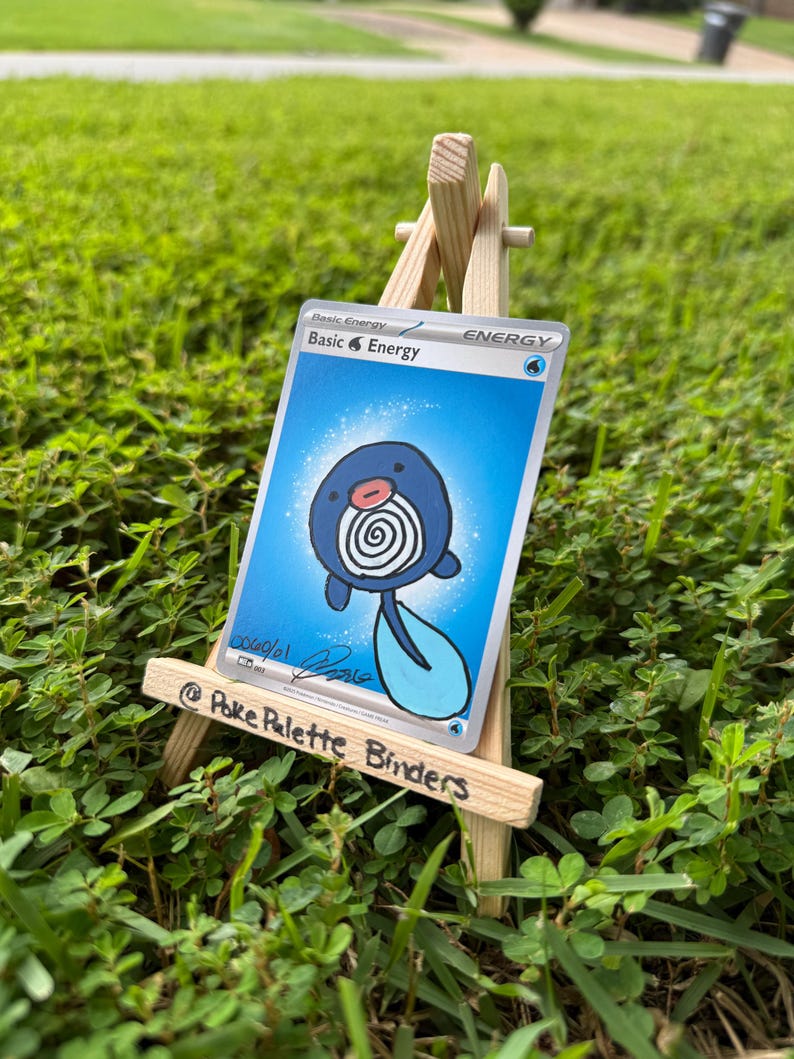 May include: A collectible trading card featuring a hand-drawn illustration of a blue Poliwag character on a Basic Water Energy card background, displayed on a small wooden easel with the text Poke Palette Binders written on the front rail.
