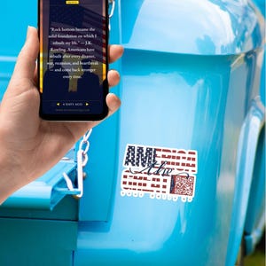 May include: A smartphone displaying a quote about resilience on its screen, held in front of a bright blue vehicle surface featuring a rectangular sticker with the text AMERICA THE GREAT, a script word The, and a QR code in red, white, and blue.