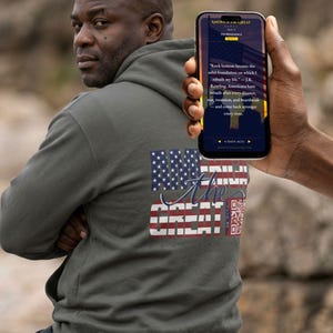 May include: An olive green hooded sweatshirt featuring a graphic on the back with the American flag and the text AMERICA THE GREAT. A smartphone is held up displaying a screen with the text AMERICA THE GREAT and a quote about resilience.