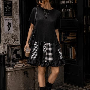 May include: A black short-sleeved dress featuring a ribbed bodice and a drop-waist skirt with a ruffled hem. The skirt is constructed from alternating panels of black fabric, white and black checkered patterns, and white and black buffalo plaid.