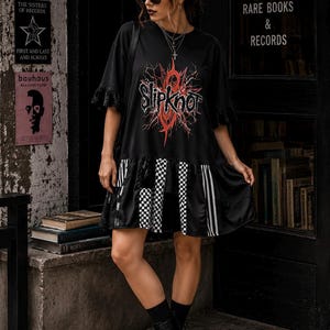 May include: A black t-shirt dress featuring a red and white Slipknot graphic on the chest. The dress has short sleeves with lace trim and a drop-waist skirt panel that alternates between black fabric, white and black checkered patterns, and vertical stripes.