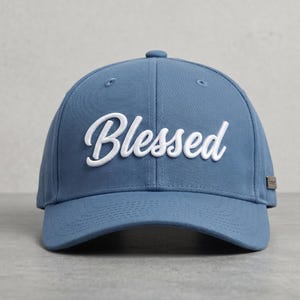 Puede incluir: Una gorra de béisbol azul polvoriento con la palabra Blessed bordada en letras cursivas blancas en los paneles frontales. La gorra tiene una corona estructurada, una visera curva y un pequeño detalle metálico rectangular en el lateral.