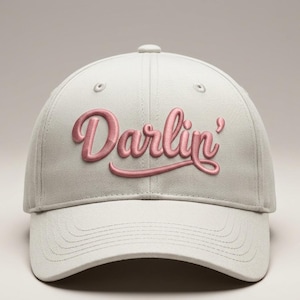 May include: A light gray baseball cap featuring the word Darlin' embroidered in a raised, metallic rose-gold script font across the front panels. The cap includes a curved brim with stitched detailing and a button at the top center.