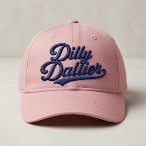 May include: A dusty rose baseball cap featuring the text Dilly Dallier embroidered in dark navy blue thread across the front panels. The cap has a structured crown, a curved brim with visible stitching, and a button at the top center.