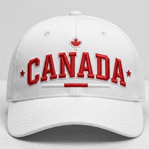 Op de afbeelding: Een witte honkbalpet met het woord CANADA in dikke rode letters op de voorkant geborduurd. Boven de tekst staat een klein rood esdoornblad en aan weerszijden van het woord CANADA staat een kleine rode ster. Onder de tekst staat een rode lijn.