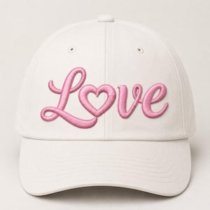 May include: A cream-coloured baseball cap featuring the word Love embroidered in a raised, puffy pink script font across the front panels. The letter o is stylised as a heart shape, and the cap includes a curved brim and stitched eyelets.
