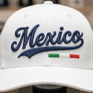 May include: A white baseball cap featuring the word Mexico embroidered in navy blue script font across the front. Below the text, a small rectangular patch displays the green, white, and red colors of the Mexican flag in horizontal stripes.