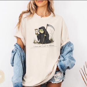 以下が含まれることがあります： アイボリー色のTシャツ。死神の格好をした緑色のカエルのグラフィックが描かれており、カエルは鎌と砂時計を持ち、足元には小さな頭蓋骨がある。イラストの下には「I know what I want for Halloween.」というテキストが記されている。