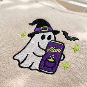 May include: A beige sweatshirt featuring an embroidered design of a white ghost wearing a black witch hat with a purple band. The ghost holds a purple can labeled Alani with a green cauldron graphic. A black bat and three green stars surround it.