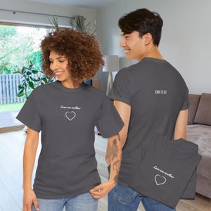 Könnte beinhalten: Ein dunkelgraues Kurzarm-T-Shirt mit dem weißen Schriftzug "Love one another" über einem einfachen weißen Herzumriss auf der Vorderseite. Die Rückseite des T-Shirts zeigt den weißen Schriftzug "JOHN 13:34" mittig am Halsausschnitt.
