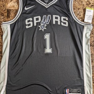 May include: A black San Antonio Spurs basketball jersey featuring the number 1 in white with grey trim. The jersey has a white Nike swoosh logo on the chest, a V-neck collar, and white side panels. A tag on the bottom left reads Engineered 52.