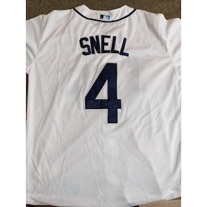 May include: A white baseball jersey featuring the name SNELL and the number 4 in dark navy blue lettering on the back. The number 4 includes a dark signature across its center. A small MLB logo patch is centered at the top of the collar.