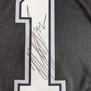 May include: A black athletic jersey featuring a large white number one with a dark grey outline. The number is screen-printed on a textured mesh fabric and displays a handwritten signature in black ink across the center of the digit.
