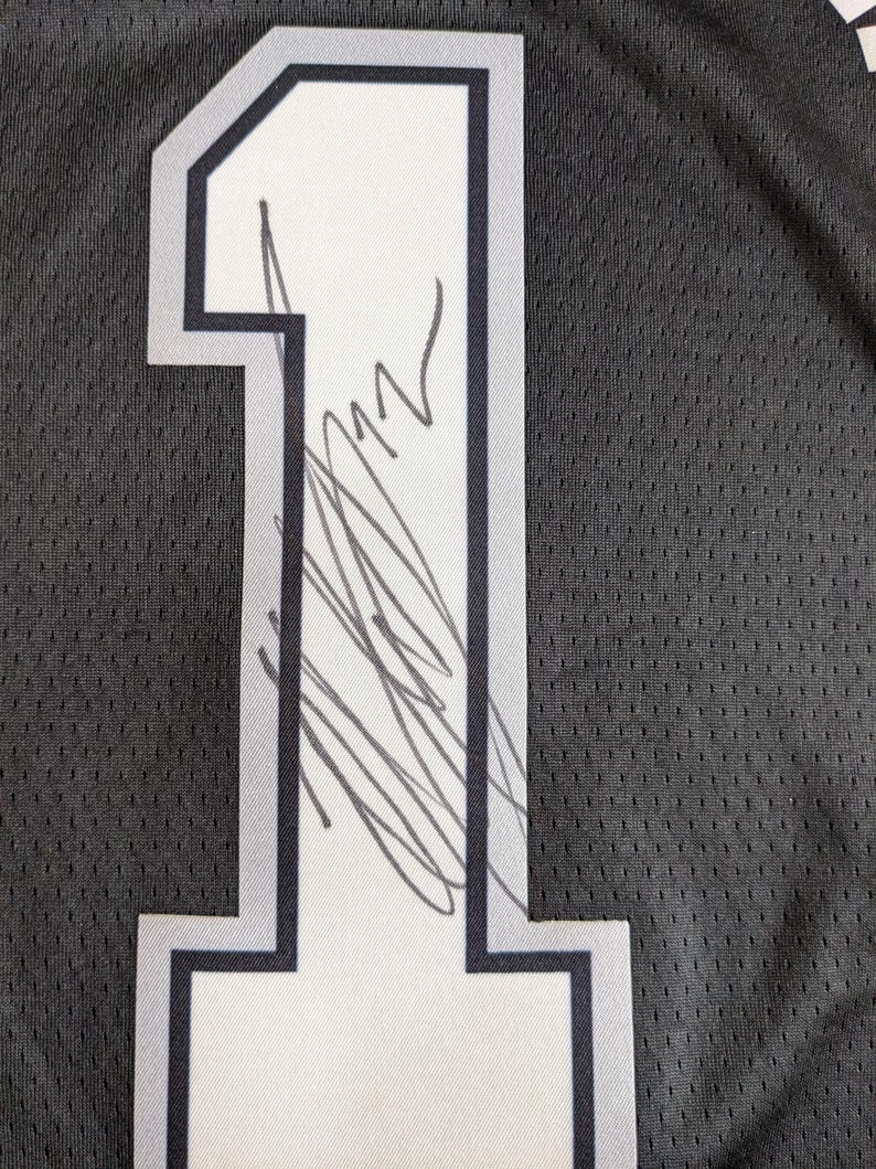May include: A black athletic jersey featuring a large white number one with a dark grey outline. The number is screen-printed on a textured mesh fabric and displays a handwritten signature in black ink across the center of the digit.