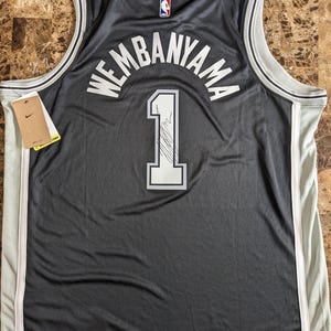 May include: A black basketball jersey featuring the name WEMBANYAMA and the number 1 in white lettering on the back. The jersey includes a small NBA logo at the neckline, white and grey trim along the armholes and collar, and a Nike brand tag attached.