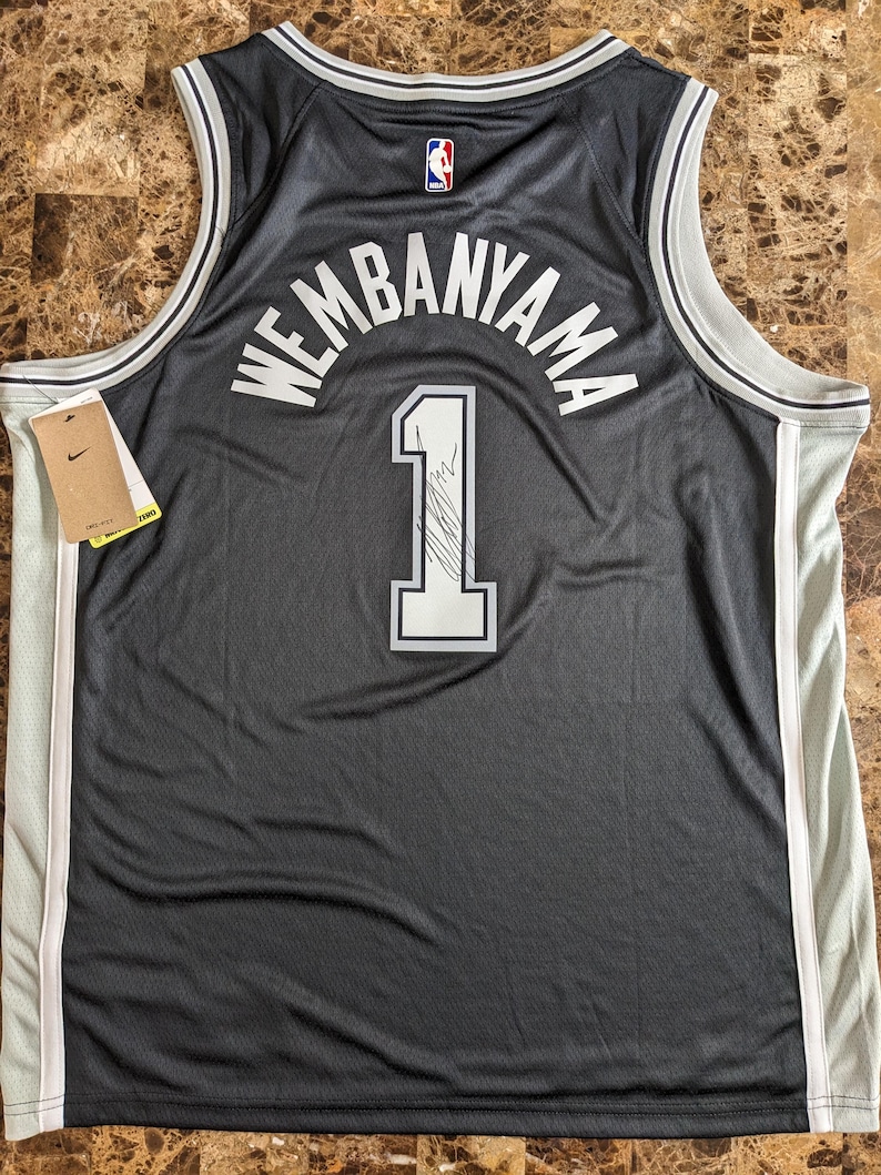 May include: A black basketball jersey featuring the name WEMBANYAMA and the number 1 in white lettering on the back. The jersey includes a small NBA logo at the neckline, white and grey trim along the armholes and collar, and a Nike brand tag attached.