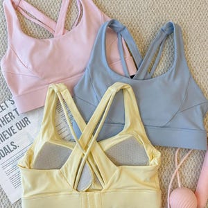 May include: Three sports bras in pastel colors: light pink, light blue, and yellow. The bras have crisscross straps on the back and a scoop neckline. A pink jump rope is visible in the background. The bras are on a textured surface.