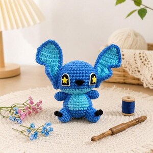 May include: A handmade blue crochet character with large ears and star-shaped eyes. The plush toy is sitting on a white, fringed mat, with a wooden crochet hook and a spool of blue thread nearby. Decorative flowers add a touch of color.