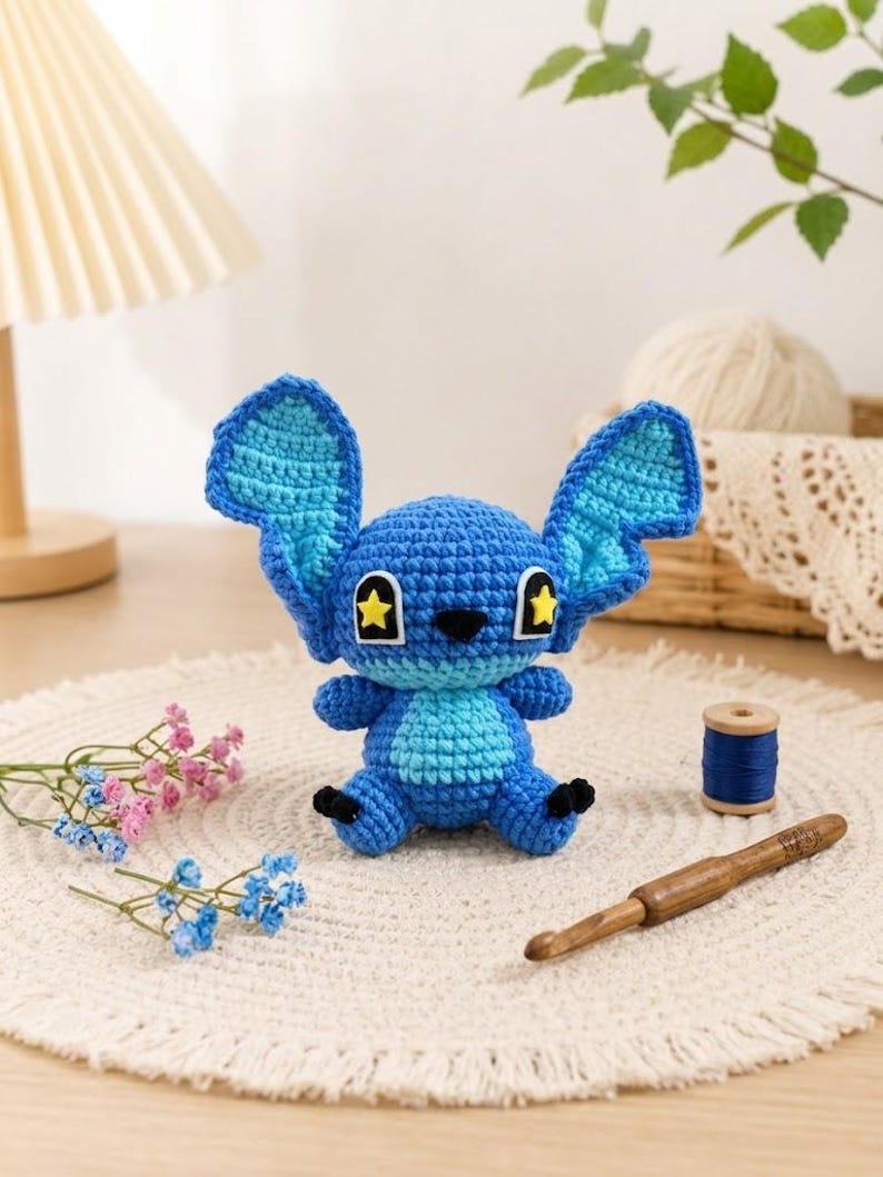 May include: A handmade blue crochet character with large ears and star-shaped eyes. The plush toy is sitting on a white, fringed mat, with a wooden crochet hook and a spool of blue thread nearby. Decorative flowers add a touch of color.
