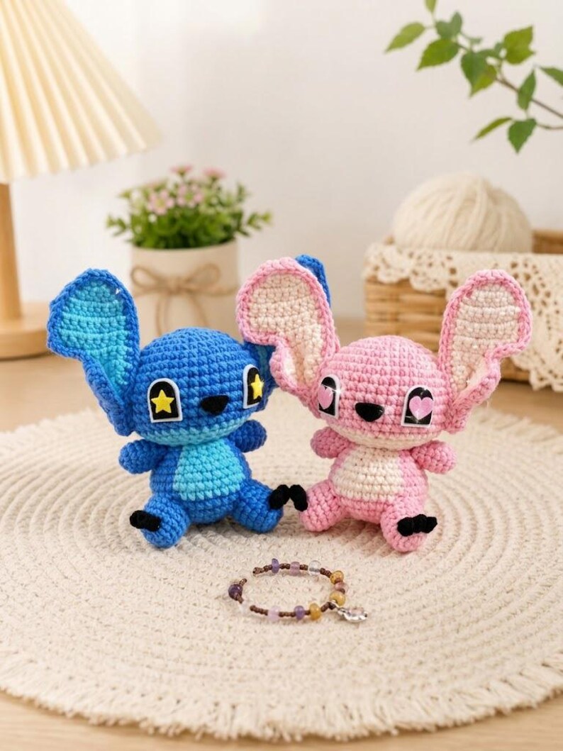 May include: Two crocheted plush toys, one blue and one pink, resembling a cartoon character. The blue toy has a yellow star on its ear, while the pink one has a heart. A beaded bracelet with a charm is in front of them. The toys are on a white, textured surface.