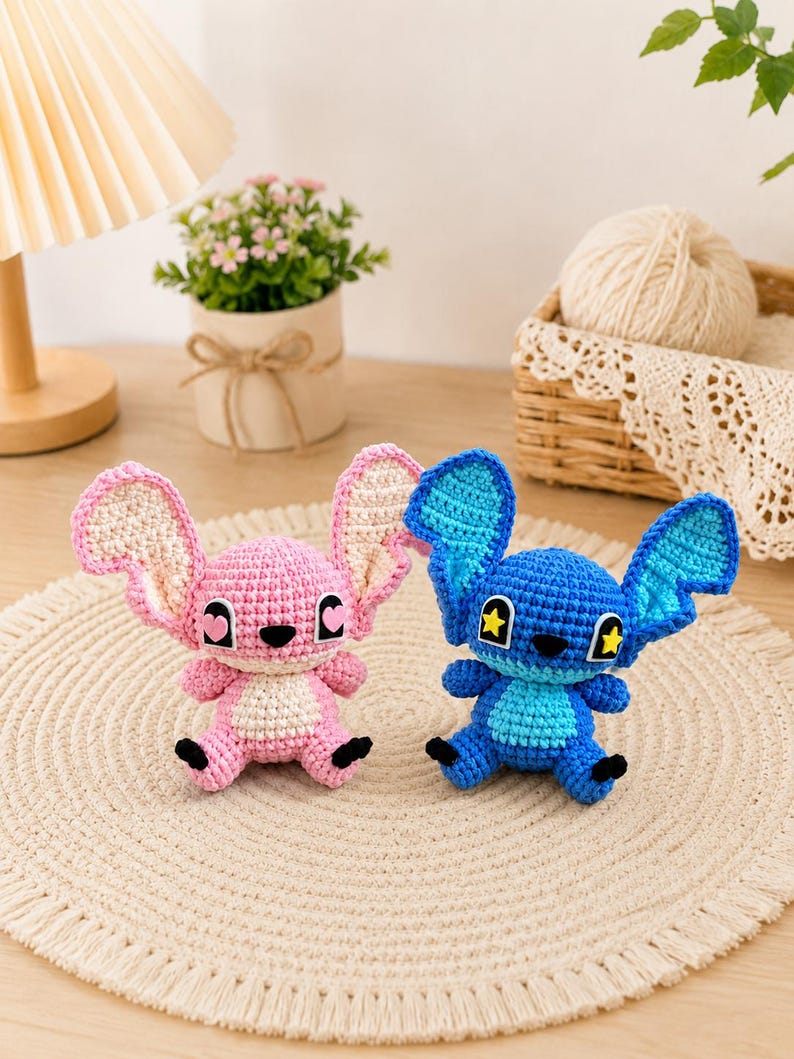 May include: Two crocheted plush toys, one pink and one blue, resembling a cartoon character. The pink toy has light pink ears and a white belly, while the blue toy has blue ears and yellow star accents. Both have black eyes and feet.