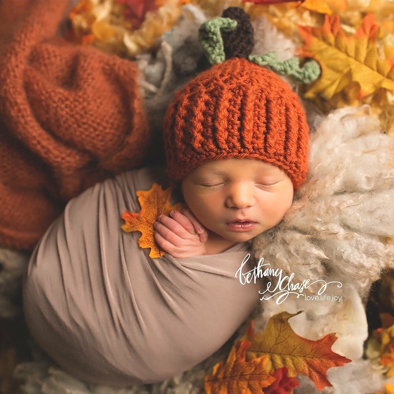 May include: A newborn baby wearing a crocheted orange pumpkin hat with green leaves, sleeping on a brown and white fluffy blanket surrounded by autumn leaves.