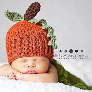 May include: A newborn baby wearing a crocheted pumpkin hat with green leaves and a brown stem. The baby is sleeping peacefully on a white blanket.