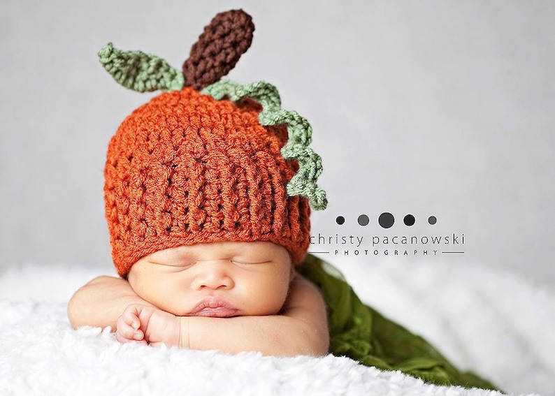 May include: A newborn baby wearing a crocheted pumpkin hat with green leaves and a brown stem. The baby is sleeping peacefully on a white blanket.