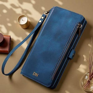 May include: A rectangular blue wallet with a wrist strap, featuring a side zipper compartment, a snap button closure, and the gold-embossed initials DN on the front. The wallet has visible stitching along the edges and a smooth, matte finish.