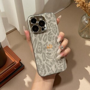 May include: A smartphone case featuring a light gray and white leopard print pattern with a metallic silver camera lens frame. The center of the case is personalized with the gold-colored letters AD. The case has a smooth, reflective finish.