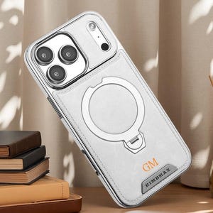 May include: A silver-toned smartphone case with a white textured back panel, featuring a circular magnetic ring stand, a camera lens cutout, and the text GM and RIMBMAX printed on the lower section of the protective cover.