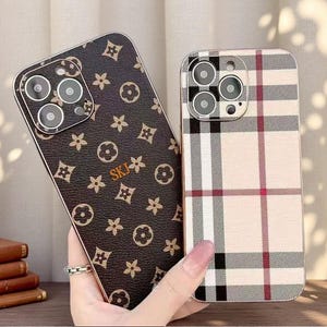 May include: Two smartphone cases with triple camera cutouts. One case features a dark brown textured surface with a repeating tan floral and geometric pattern and the letters SKJ in gold. The other case has a beige, black, and red plaid pattern.