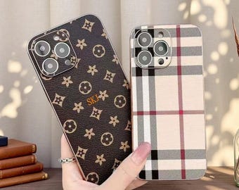 May include: Two smartphone cases with triple camera cutouts. One case features a dark brown textured surface with a repeating tan floral and geometric pattern and the letters SKJ in gold. The other case has a beige, black, and red plaid pattern.