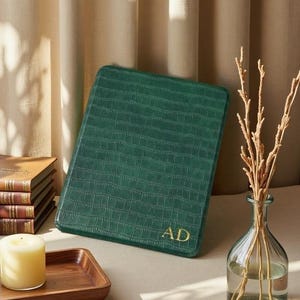 May include: A green journal with a crocodile skin effect and gold initials "AD". The journal is rectangular and is displayed with a candle, books, and a vase with dried stems. The journal is likely A5 size, approximately 14.8 x 21 cm.
