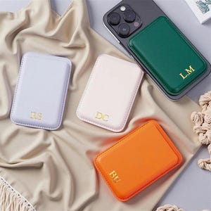 Op de afbeelding: Een assortiment rechthoekige telefoonportemonnees in pastelkleuren zoals lavendel, roze en oranje, plus een groene portemonnee bevestigd aan een telefoon. Elke portemonnee heeft gouden initialen. De portemonnees zijn gerangschikt op een neutraal gekleurde stof.