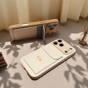 May include: Two beige and off-white phone cases with cardholder pockets. One case is open, revealing the cardholder. The other case is closed. The initials "LM" are printed on the cardholder of the off-white case.