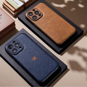 Könnte beinhalten: Zwei rechteckige Smartphone-Hüllen mit strukturierter Oberfläche und erhöhten Kameraaussparungen. Eine Hülle ist hellbraun, die andere marineblau mit den mittig eingeprägten Buchstaben GM. Beide Hüllen haben genähte Kanten und ein schmales Design.