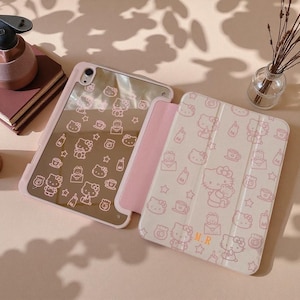 Könnte beinhalten: Eine Tablethülle mit einer reflektierenden Rückseite und einem hellrosa Klappdeckel, beide verziert mit einem wiederkehrenden Muster aus Hello Kitty, Sternen und Kaffeetassen. Der vordere Deckel zeigt die orange gestickten Initialen M.R.