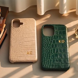 Könnte beinhalten: Zwei Handyhüllen mit Krokodilhautstruktur. Eine ist beige mit den Initialen "AC" in Gold, die andere ist dunkelgrün mit den Initialen "KD" in Gold. Die Hüllen befinden sich auf einem neutralen Hintergrund.