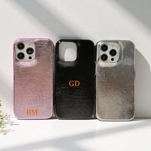 Puede incluir: Tres fundas de teléfono personalizadas en rosa, negro y plateado. La funda rosa tiene las iniciales "BM", la funda negra "GD" y la funda plateada está en blanco. Todas las fundas tienen un acabado metálico texturizado.