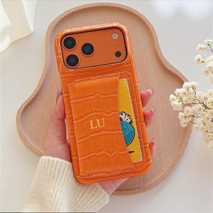 Puede incluir: Funda de teléfono naranja con textura de cocodrilo y tarjetero. El tarjetero tiene las iniciales "LU" en relieve. Una tarjeta amarilla y azul es parcialmente visible.
