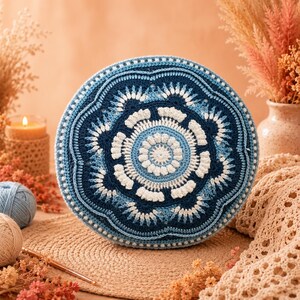 Könnte beinhalten: Ein rundes Häkelkissen mit einem Mandala-Muster in Marineblau, Hellblau und Weiß. Das Design besteht aus konzentrischen Kreisen mit strukturierten Maschen, einschließlich Noppen und Muscheln, die ein blumenartiges geometrisches Motiv bilden.