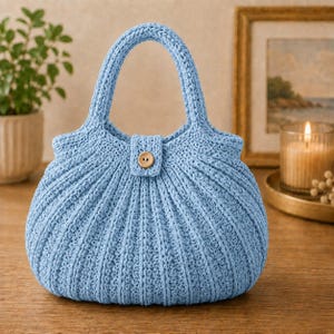 Könnte beinhalten: Eine hellblaue gehäkelte Handtasche mit strukturiertem Rippenmuster, einem einzelnen abgerundeten Griff und einem mittigen Laschenverschluss, der mit einem kleinen, runden, hellbraunen Holzknopf befestigt ist. Die Tasche hat eine geschwungene Form.
