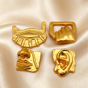 May include: A set of four gold-toned metal decorative pieces featuring anatomical shapes. The collection includes a stylized eye, a hand forming a fist, a nose with lips, and an ear. Each piece has a smooth, reflective surface and irregular edges.