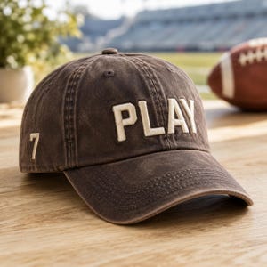 May include: A brown baseball cap with a distressed finish featuring the word PLAY embroidered in large cream-colored letters across the front panels and the number 7 embroidered in the same cream color on the side panel of the cap.