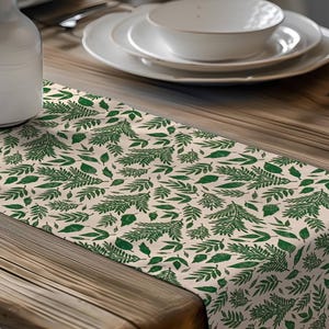 May include: A beige table runner with a repeating pattern of green leaves and ferns. The runner is placed on a wooden table, with white dishes and a white vase with dried flowers in the background. The runner adds a natural, botanical touch to the table setting.